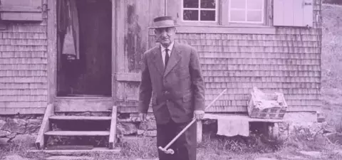 An elderly man in a suit and hat stands with a cane outside a rustic wooden cabin, with a stone path and basket nearby.