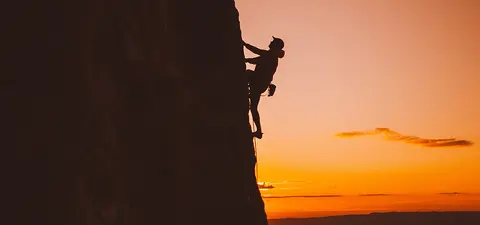 A lone rock climber grips a steep cliff in silhouette against a glowing orange sunset, creating a dramatic, adventurous scene of courage and solitude.