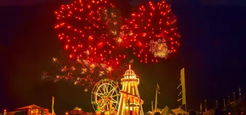 A vibrant nighttime scene featuring fireworks illuminating the sky above a fairground, with a ferris wheel and festive structures in the foreground.