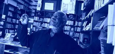 An older man in a dark shirt stands in a library, gesturing upward while holding glasses, surrounded by bookshelves filled with books.