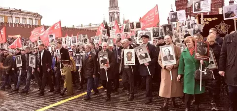 A large crowd marches in a parade, holding portraits of historical figures, with flags in the background, near the Kremlin in Moscow.