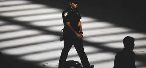 A police officer walks through a space lit by striped shadows, with another figure partially visible in the darkness nearby.