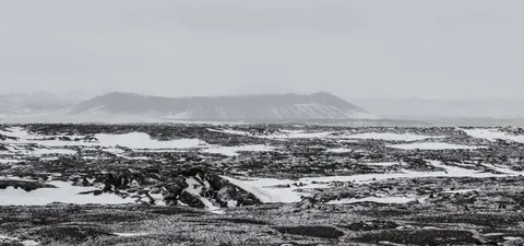 A barren, snowy landscape with dark volcanic rock formations under a cloudy sky, featuring distant mountains and a sense of isolation.