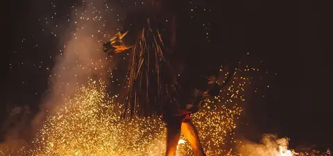 A person wearing traditional attire walks through flying sparks and flames, creating a dramatic scene illuminated by firelight in a dark setting.
