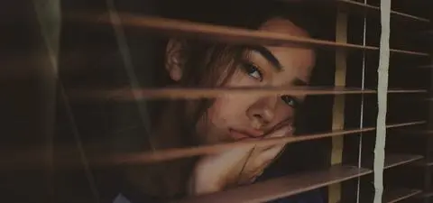 A contemplative young woman gazes through horizontal wooden blinds, resting her chin on her hand with a thoughtful expression.