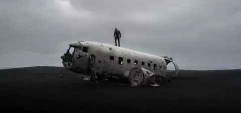 A lone figure stands atop a wrecked airplane on a desolate, dark landscape under a cloudy sky.