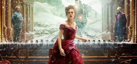A woman in a red gown stands on a snowy train platform, with a vintage train in the background and two men observing her.