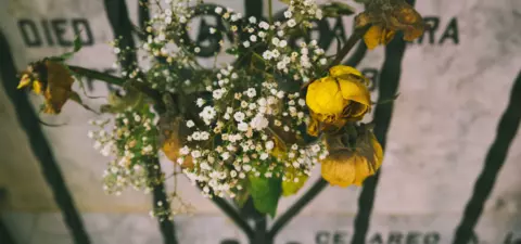 A bouquet of dried flowers, including yellow roses and small white blooms, placed against a weathered gravestone with inscriptions.
