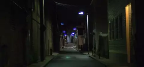 A dark, narrow alley illuminated by street lamps, featuring brick buildings on either side and a faint light at the end of the path.