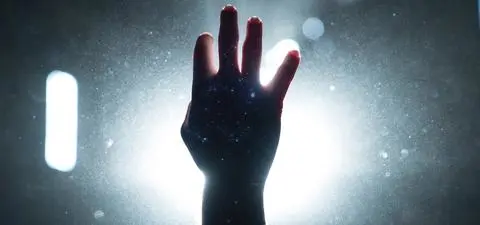 A silhouette of a raised hand against a bright light source, with scattered particles creating a dramatic atmosphere.