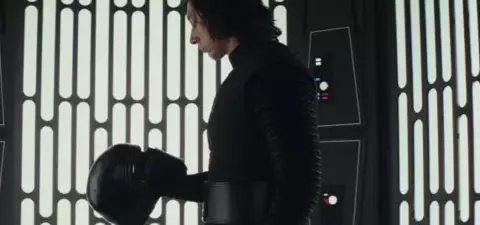 A hooded figure in a dark suit stands in a futuristic setting, holding a black helmet, with vertical light patterns in the background.