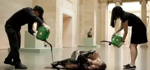 Performance art piece featuring two individuals pouring black liquid over a person lying on the floor in a museum setting, emphasizing themes of vulnerability and transformation.