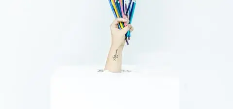 A hand emerges from a white box, holding a colorful array of pencils, against a plain white background.
