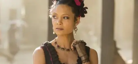 A woman with curly hair adorned with flowers, wearing a vintage-style dress and jewelry, stands thoughtfully against a soft, blurred background.