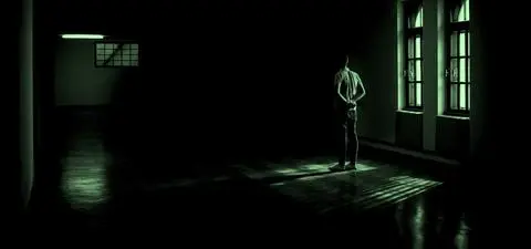 A solitary figure stands in a dimly lit room, gazing toward windows, with shadows cast across the polished floor.