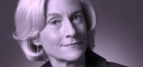 A black and white portrait of a woman with shoulder-length blonde hair, wearing a dark blazer, smiling gently while looking slightly to the side.
