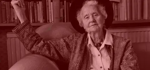 An elderly woman in a patterned jacket and hat, seated on a couch, with bookshelves filled with books in the background.