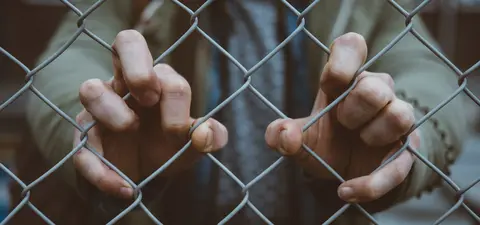 A close-up of hands gripping a chain-link fence, symbolizing confinement and longing for freedom.