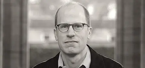 A man with glasses and a serious expression stands in front of a blurred background, wearing a black jacket over a collared shirt.