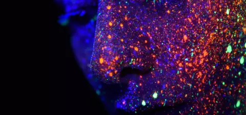 A close-up of a face adorned with vibrant neon glitter and sparkles, illuminated under ultraviolet light, creating a cosmic and otherworldly effect.