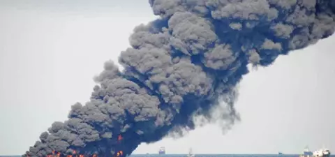 A large plume of thick black smoke rises from a marine oil fire, with flames visible at the water's surface and several ships nearby.