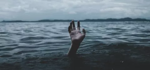A submerged hand reaches up from dark waters, with a cloudy sky and distant hills creating a dramatic backdrop.