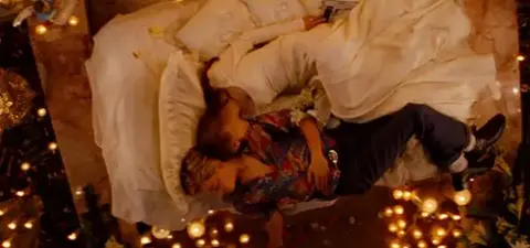 A couple in formal attire lies together on a lavish bed surrounded by candles and flowers, conveying a romantic atmosphere.