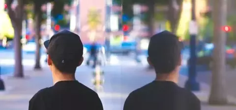 A child in a black cap stands on a sidewalk, facing a reflective surface that shows their silhouette, surrounded by urban scenery and soft greenery.
