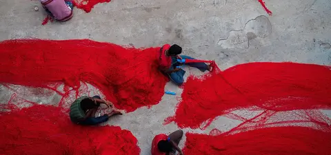 Workers are seen creating vibrant red fishing nets in a workshop, surrounded by long strands of red thread on a concrete floor.