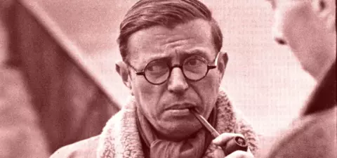 Philosopher Jean-Paul Sartre, wearing glasses and a coat with a fur collar, ponders while holding a pipe, possibly engaged in conversation with another person.