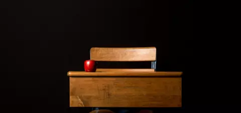 A wooden school desk with a chair, featuring a red apple on top, against a black background.