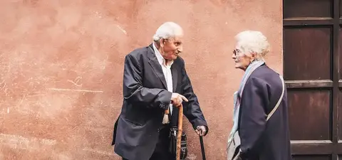 Two elderly individuals converse on a street, with one using a cane and both dressed in stylish attire, against a textured pink wall.