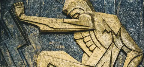 A golden relief sculpture of a worker in a dynamic pose, showcasing muscular arms and industrial elements, representing strength and perseverance.