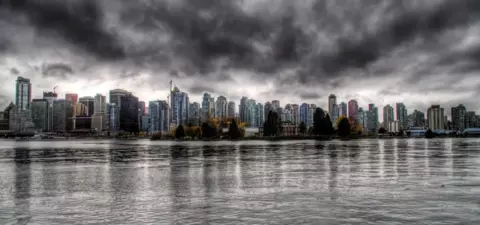 A dramatic skyline of Vancouver reflects on the water, with tall buildings under a cloud-covered sky, creating a moody atmosphere.