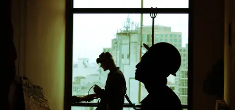 Two silhouettes of workers inside a building, with one operating equipment near a window, while the other is in profile, against a backdrop of a city skyline.