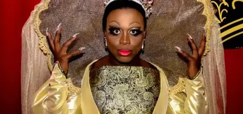 A performer in a lavish golden costume with an oversized decorative collar, adorned with a crown and striking makeup, posing against a vibrant backdrop.