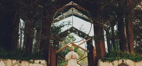 A person in a white robe stands before a striking mirrored entrance surrounded by lush greenery, creating a serene and reflective atmosphere.