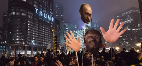 Protesters hold large cutouts of Donald Trump and Vladimir Putin while demonstrating at night in Chicago, with the Trump Tower illuminated in the background.