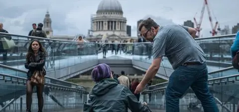 A man offers assistance to a seated figure on the Millennium Bridge, with St. Paul's Cathedral visible in the background.
