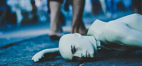 A white, lifelike mannequin lies face down on the ground, surrounded by blurred figures of people walking in a busy outdoor setting.