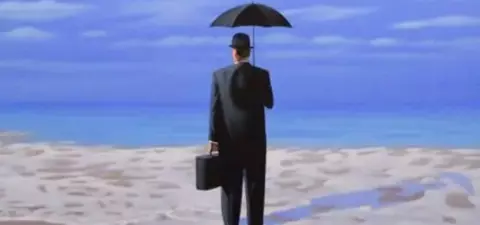 A man in a suit holding an umbrella stands on the beach, facing the ocean with a briefcase beside him, against a serene sky.