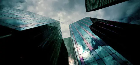 A dramatic view of tall glass skyscrapers reflecting clouds and sky, creating a dynamic urban skyline against a moody atmosphere.