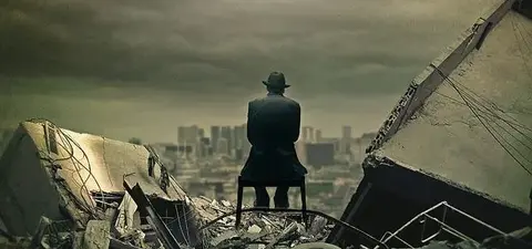 A man in a hat sits on a chair among rubble, looking out over a city skyline under a dark, cloudy sky.