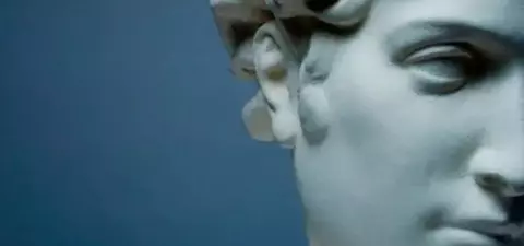 Close-up of a white marble sculpture, possibly representing a classical figure, showcasing detailed facial features and curly hair against a blue background.