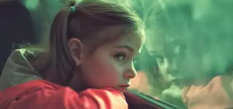 A young girl with a ponytail leans on her arms, gazing pensively out of a train window, reflecting a sense of contemplation and wonder.