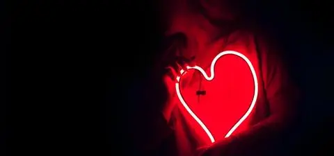 A person holds a glowing red neon heart against a dark background, creating a dramatic and romantic atmosphere.
