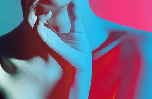 A close-up of a person's neck and hand, featuring artistic blue and pink lighting, highlighting the contours and skin texture.