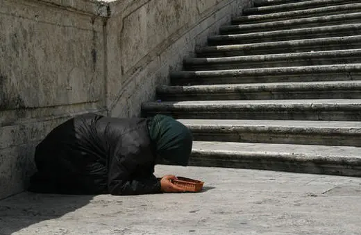 A person in a dark cloak and green headscarf kneels on a stone surface, holding a basket, beside a set of stairs.