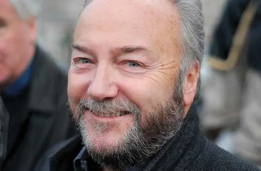 A smiling man with a beard, wearing a dark coat, stands outdoors with blurred figures in the background.