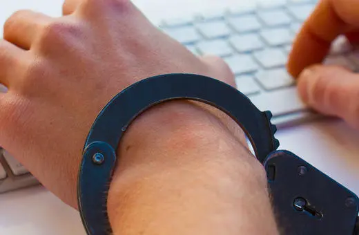 A handcuffed hand typing on a computer keyboard, symbolizing restrictions on freedom, privacy concerns, or cybersecurity issues.
